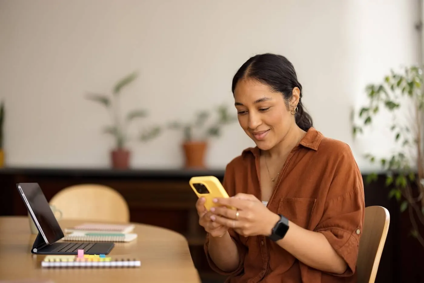 Smiling woman sitting at a desk checking a phone, with a tablet and notepads beside her