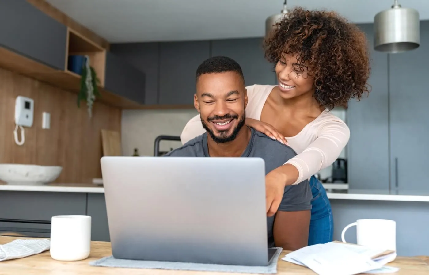 Happy couple looking at a laptop together in a kitchen, with the woman leaning over and pointing at the screen.
