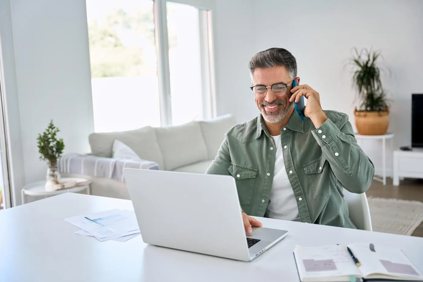 Smiling mature man sits at a desk working on a laptop while holding a phone to their ear, with papers and a notebook spread out nearby in a bright, modern living space.