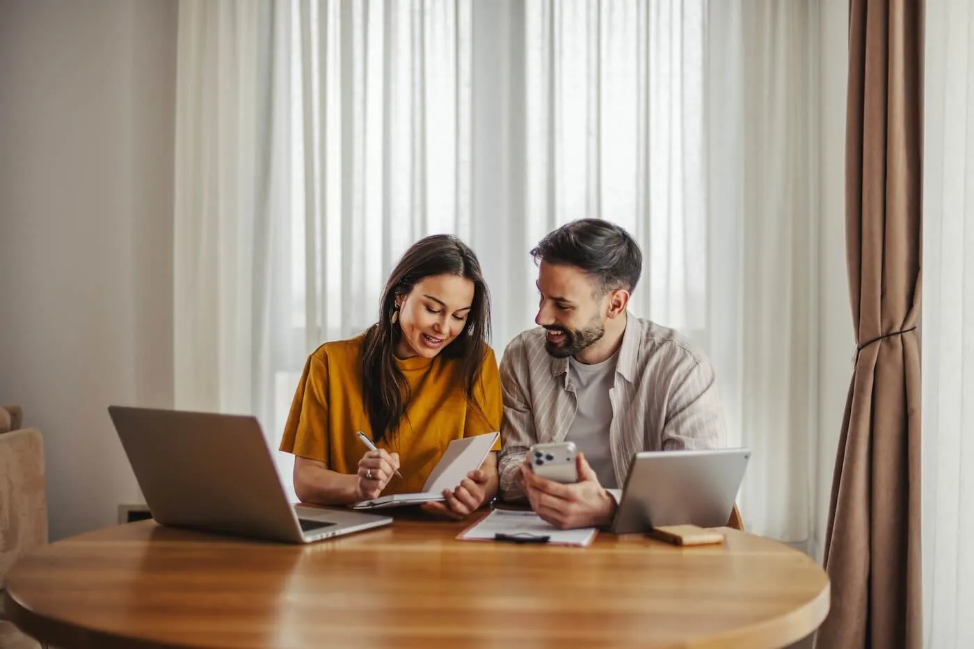 Happy young couple sitting at a round table using a laptop, smartphone, and documents while reviewing information together in a bright room.
