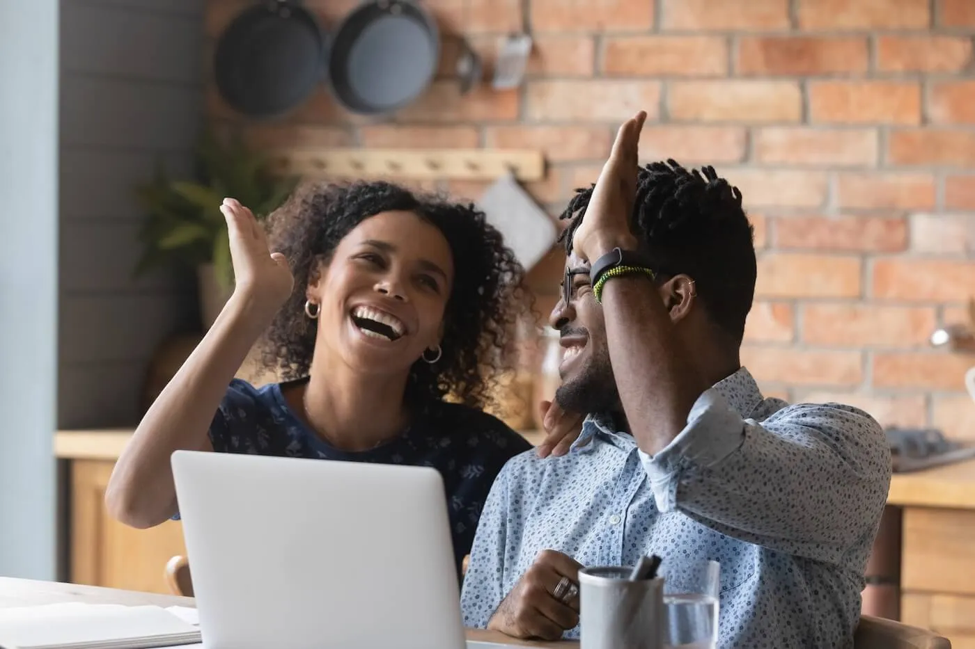 Happy couple sitting together at a table in a kitchen, giving each other a high‑five in front of an open laptop