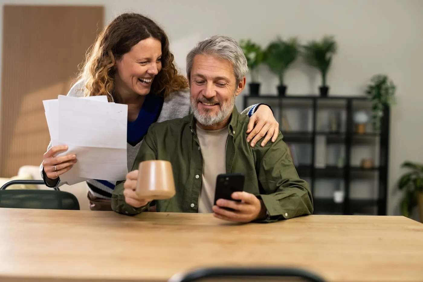Mature couple sitting at a table, with the woman holding papers and the man holding a phone and a mug, in a room with shelves and plants in the background
