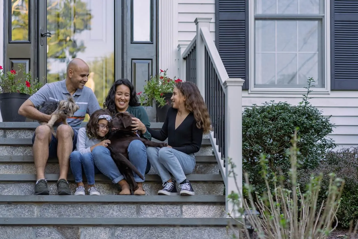 A happy family of four eated on the front steps of a house, accompanied by two dogs, with potted plants, railings, and house siding visible around the entryway.