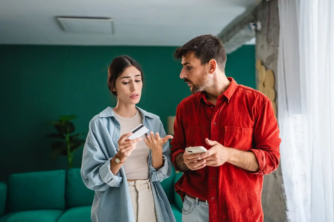 Concerned couple standing in a modern living room, holding a credit card and a mobile phone while discussing finances