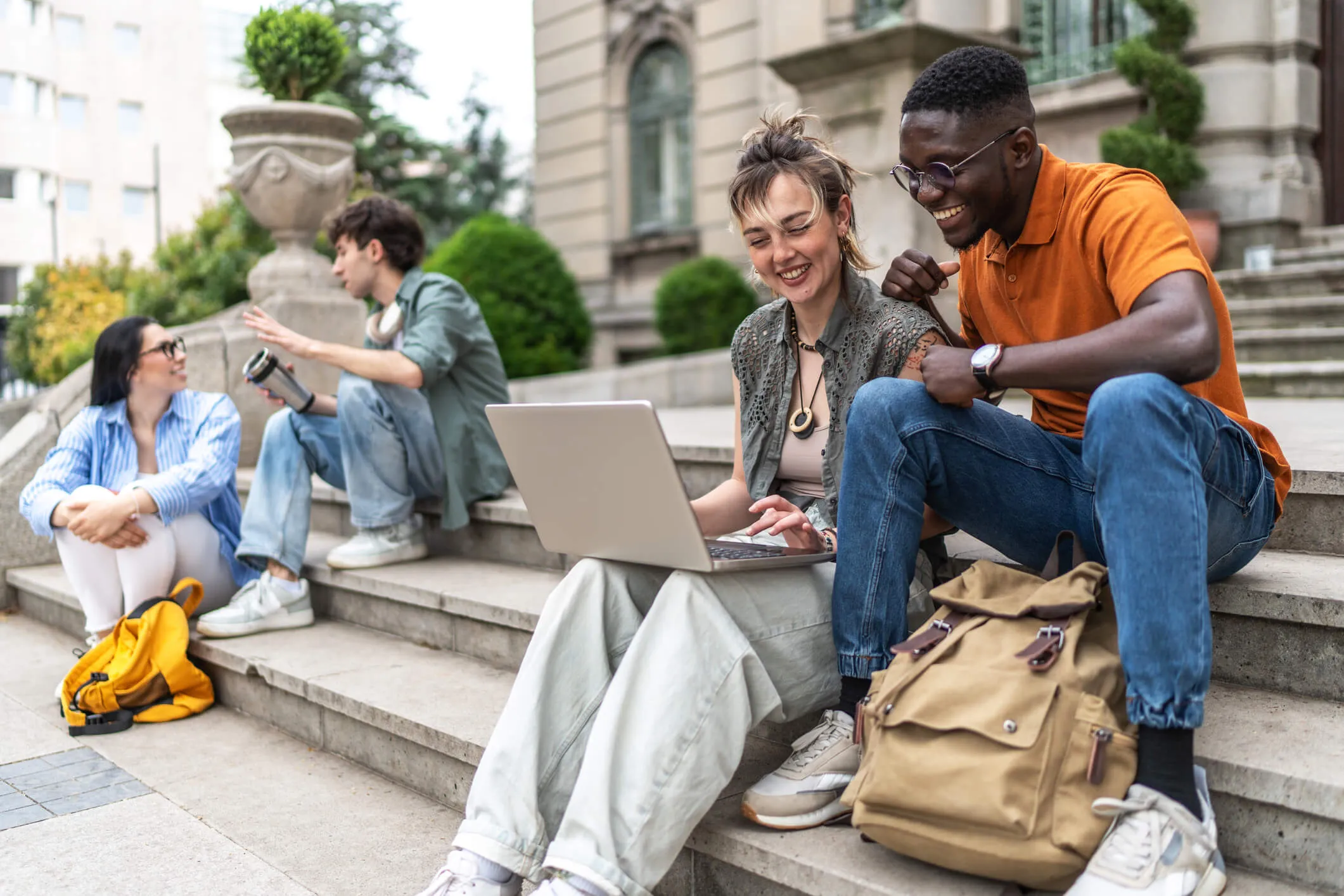 A group of college students sitting on outdoor steps using a laptop together, with backpacks nearby and a building in the background.