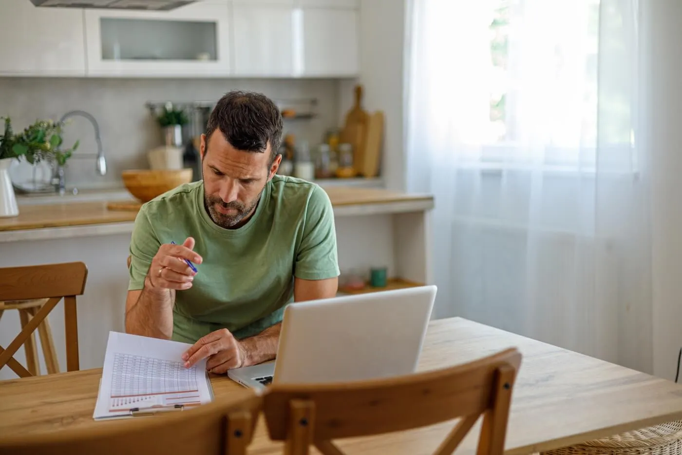 Focused mature man planning finances at the kitchen table, with an open laptop beside him