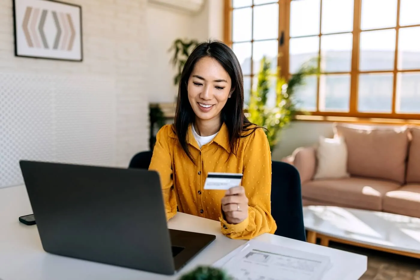 Smiling young woman sitting at a desk holding a credit card while using a laptop, with documents on the table and a bright living room in the background