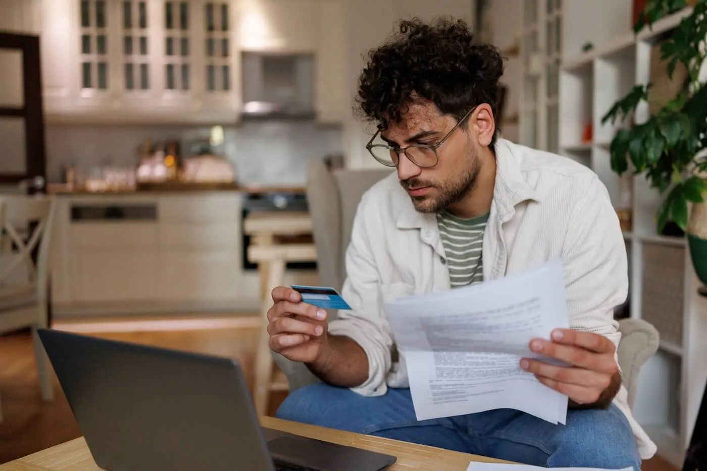 Focused man looking at his credit card while holding a printed document, with an open laptop beside him on a coffee table