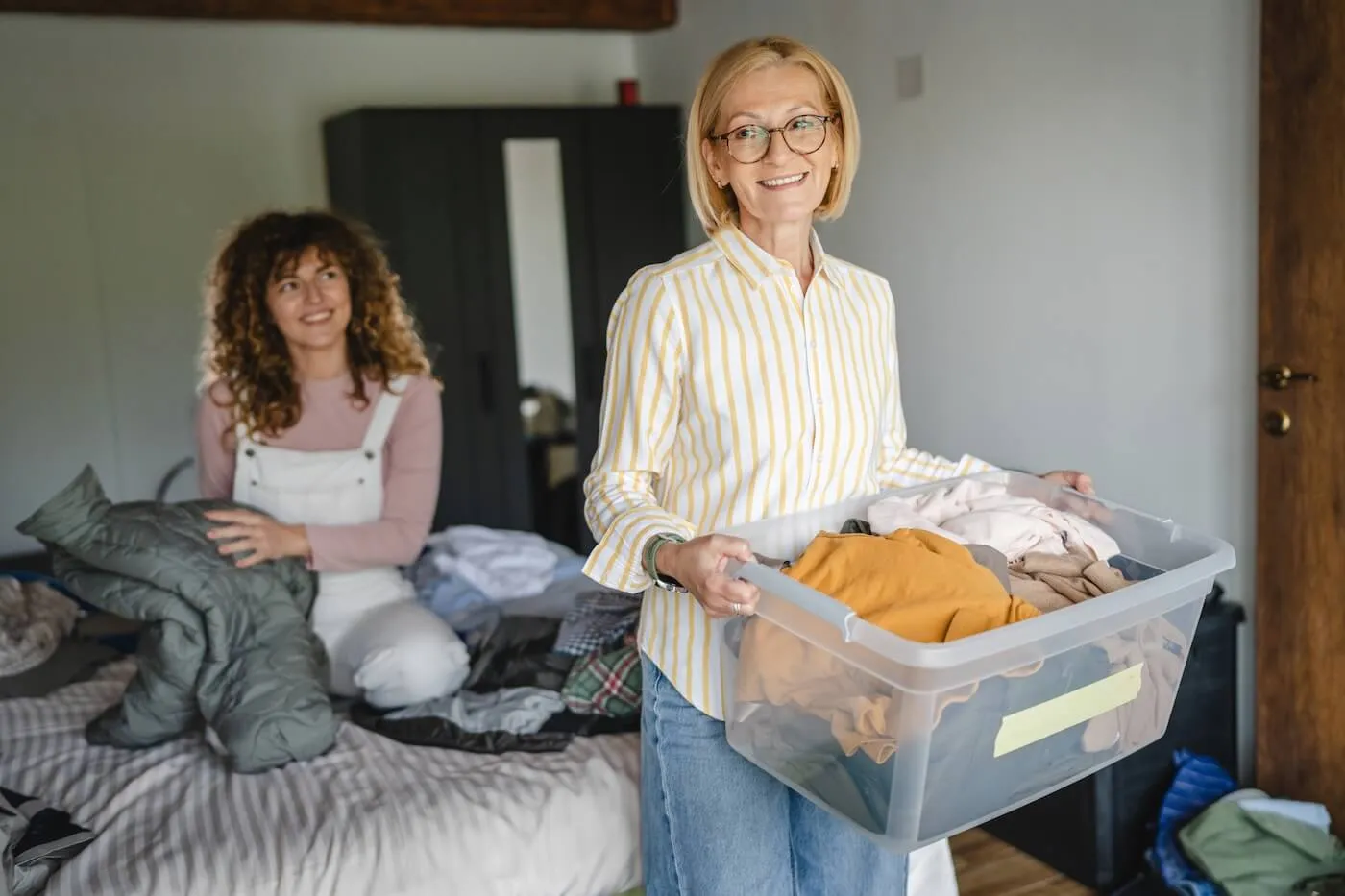 Smiling mature woman holds a plastic storage bin filled with folded clothes for donation while a younger woman sits on a bed sorting garments, in a bedroom with folded and scattered clothing.
