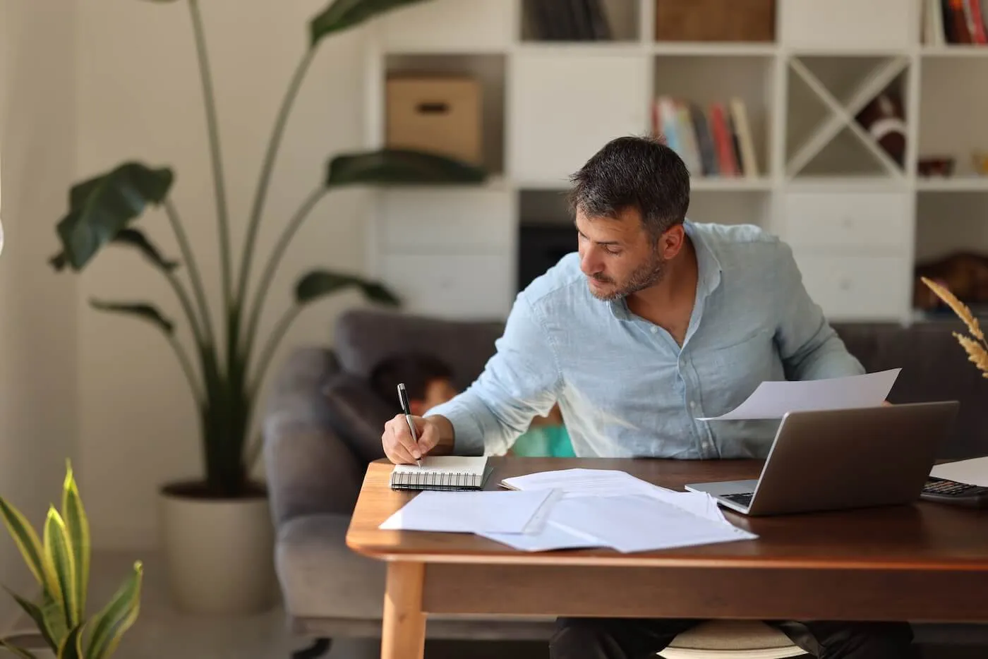 Focused man sits at a table writing on a notepad while reviewing documents, with a laptop and papers spread out in a home setting.