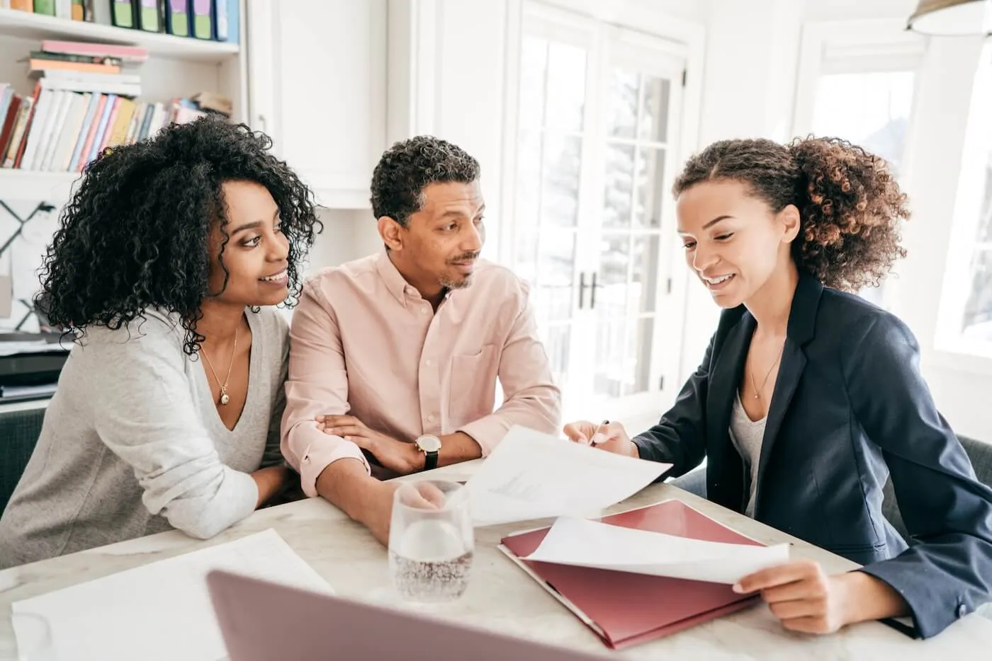 A couple and a female financial advisor seated at a bright home workspace, reviewing documents together across a table with folders, papers, and a glass of water.