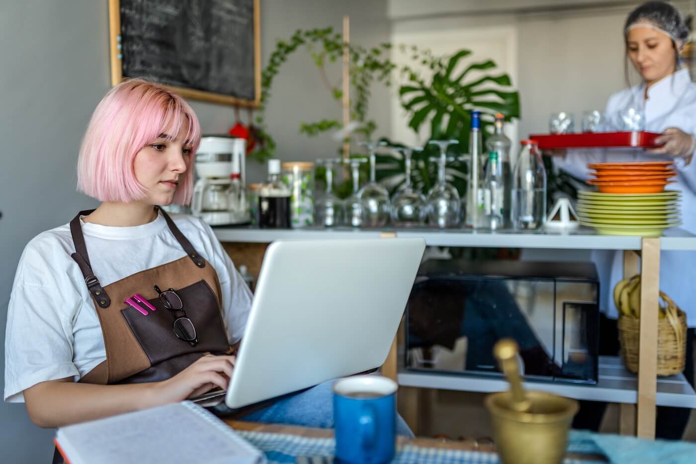 Focused young woman with pink hair wearing an apron is using laptop during her break from work in the cafe