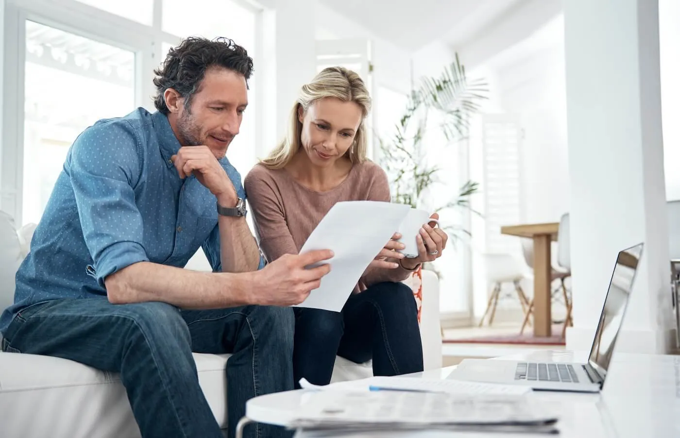 Mature couple sitting together on a couch reviewing documents, with a laptop and additional papers spread out on the table in front of them.