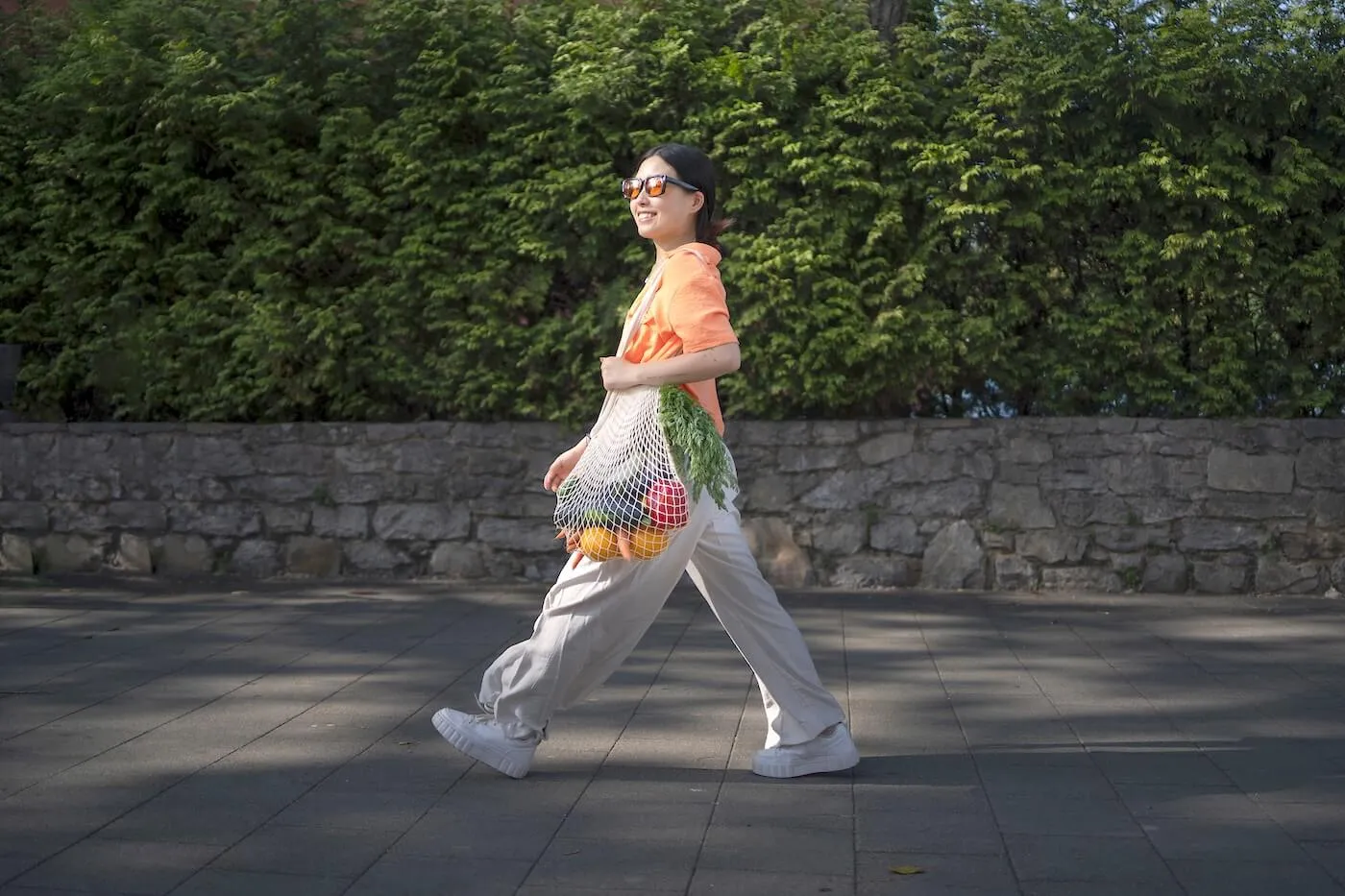 Smiling young woman walking while carrying a reusable tote bag filled with vegetables and fresh herbs