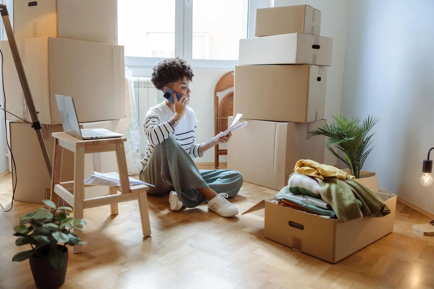 Focused woman sitting on a wooden floor surrounded by stacked cardboard moving boxes, holding papers and talking on a phone, with a laptop on a small stool and a box of folded clothes nearby in a bright room.