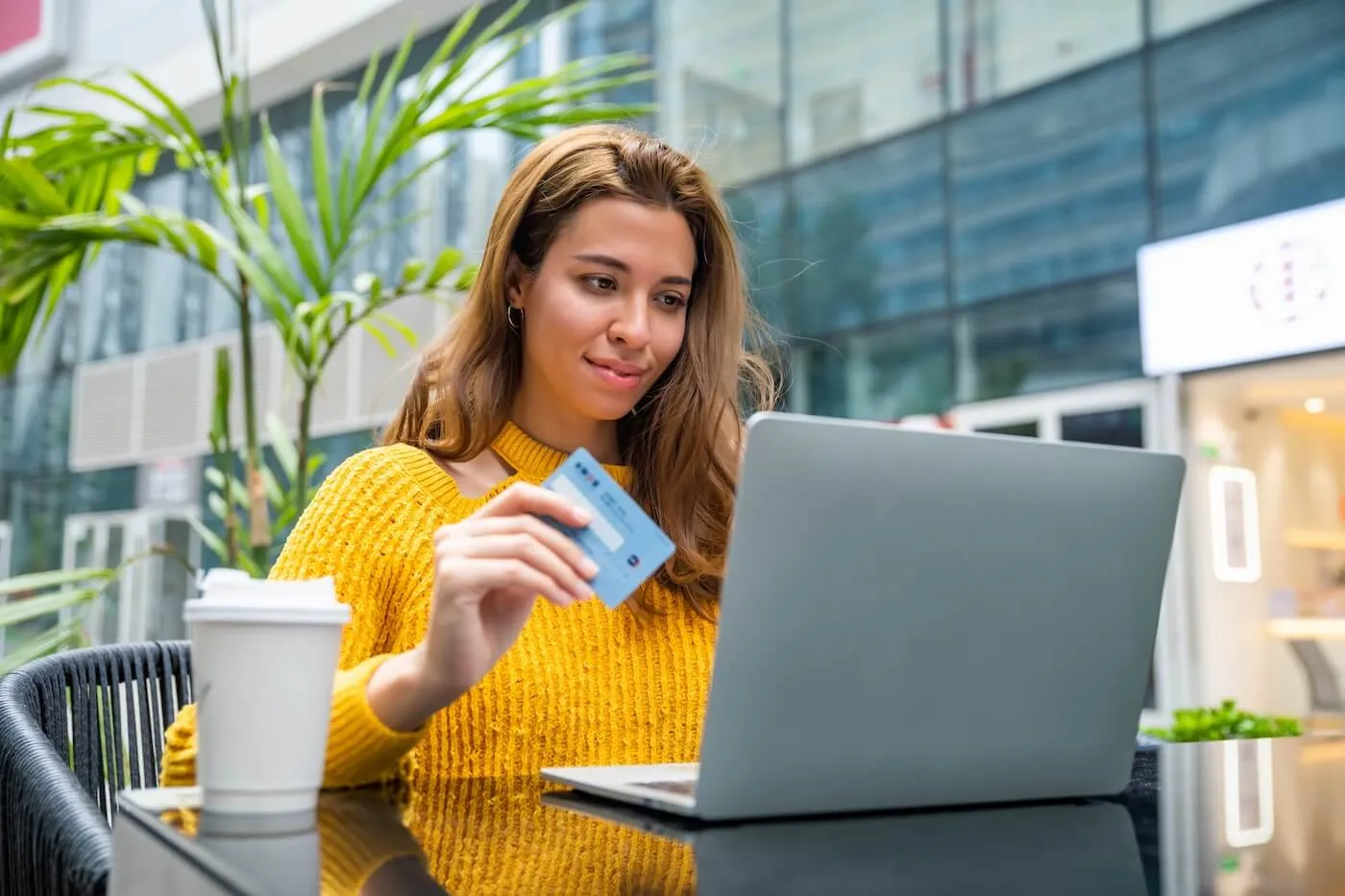 Smiling woman using her credit card to make an online payment while sitting at an outdoor café with a laptop and a paper cup beside her