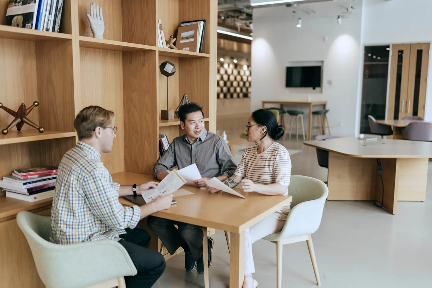 Mortgage broker seated on the left at a table meeting with a couple, reviewing documents together in a modern office with wooden shelving, books, and shared workspaces in the background