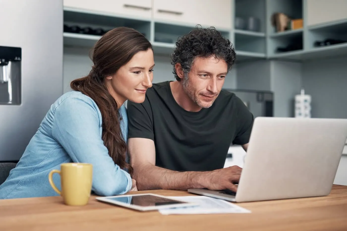 Focused couple using laptop on the kitchen counter, with a tablet, printed documents, and a mug beside them