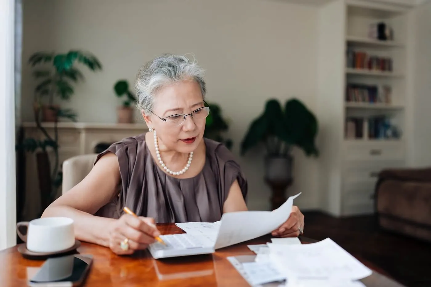 Focused senior woman sitting at a wooden table reviewing printed documents, holding a pencil and a sheet of paper, with additional papers, a calculator, a cup, and a smartphone on the table.
