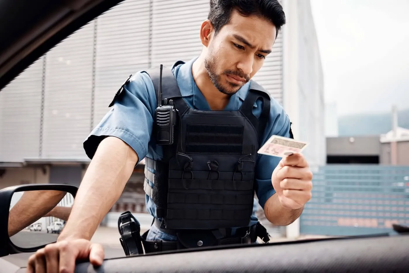 Uniformed law enforcement officer wearing protective gear and a radio, leaning toward a vehicle window while inspecting a driver's license or identification card. Office buildings and an urban streetscape are visible in the background.