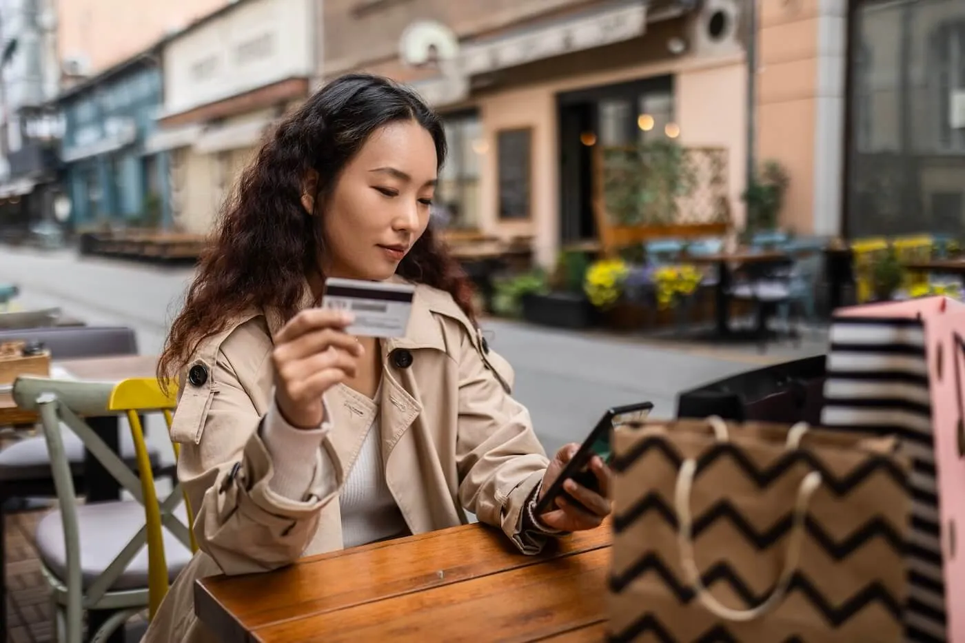 Focused woman at an outdoor café holds a credit card while using a smartphone; shopping bags sit on the table nearby.