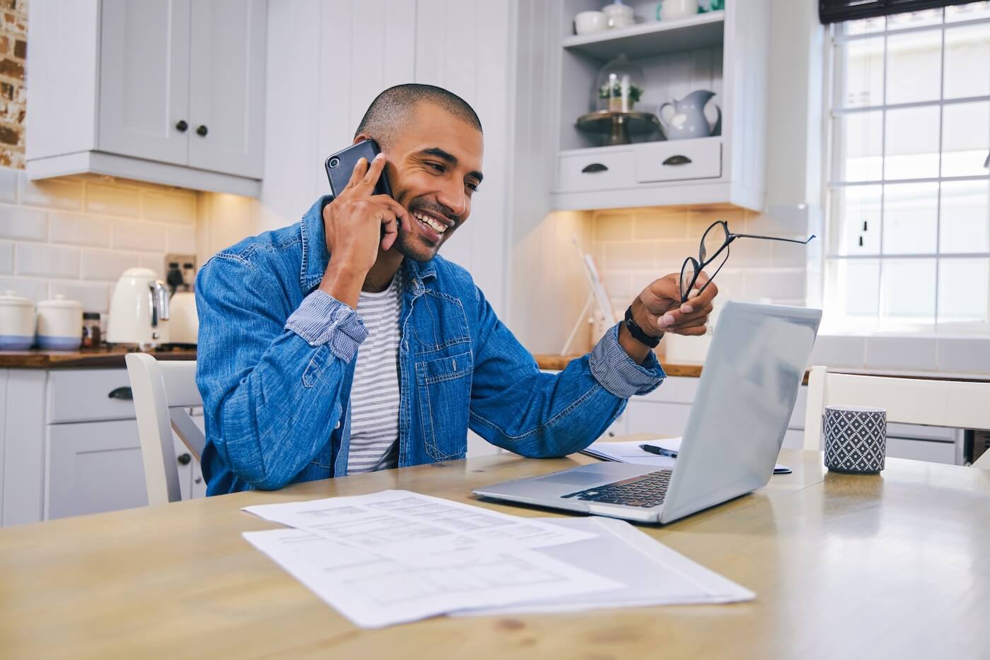 Smiling man having a phone call in the kitchen with an open laptop and printouts on the table beside him