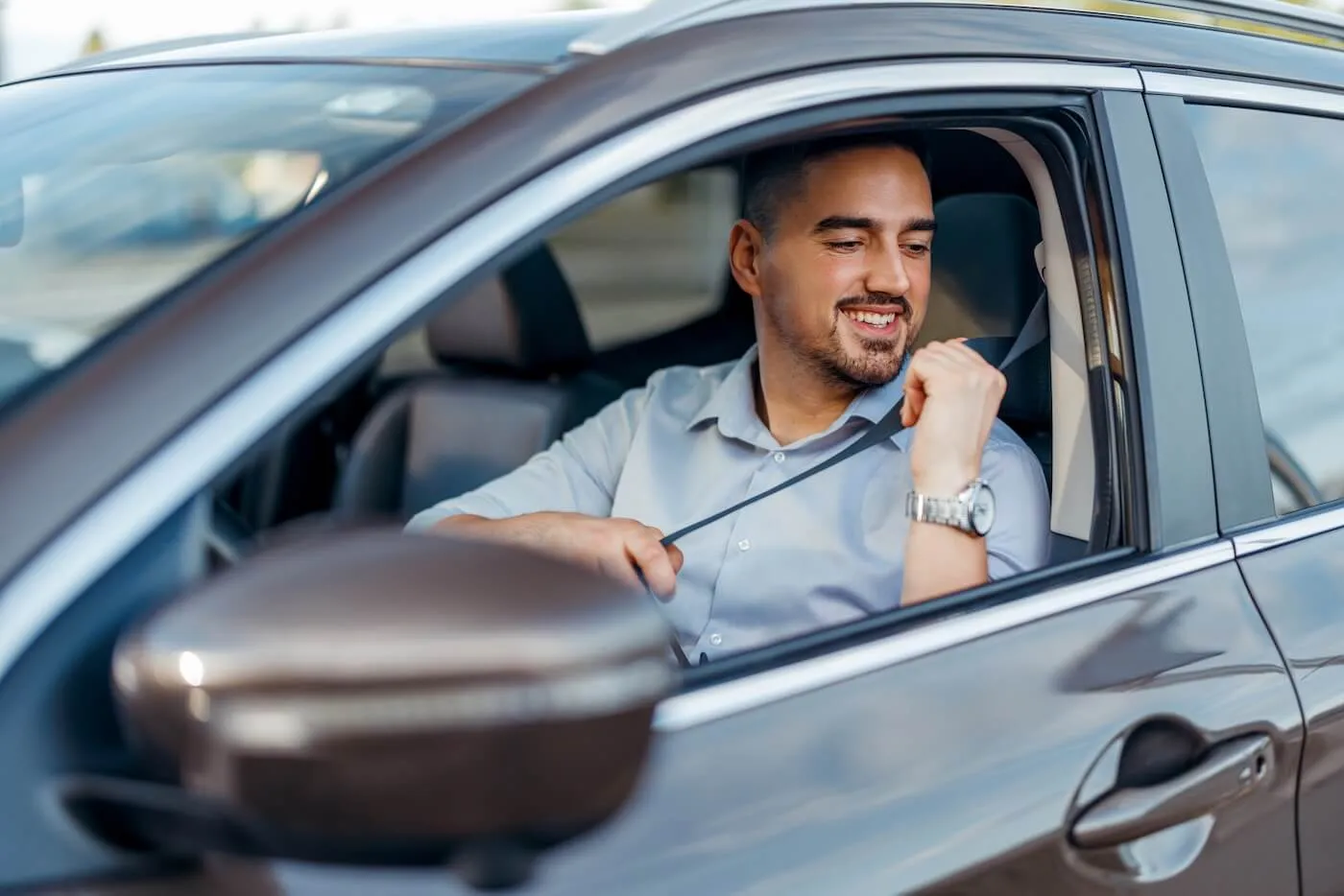 Smiling man seated in the driver's seat of a vehicle fastening a seat belt before driving. The image shows the interior of a modern car, including the side mirror, door frame, and front cabin area.