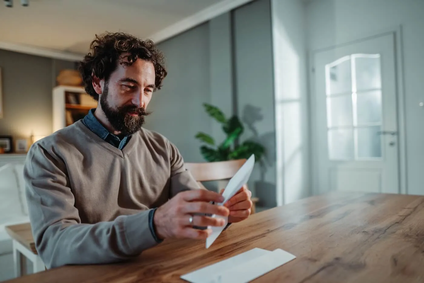 Focused man seated at a wooden dining table in a home setting, opening and reviewing a piece of mail or a document. A second envelope rests on the table, with bookshelves, a houseplant, and soft natural light visible in the background.
