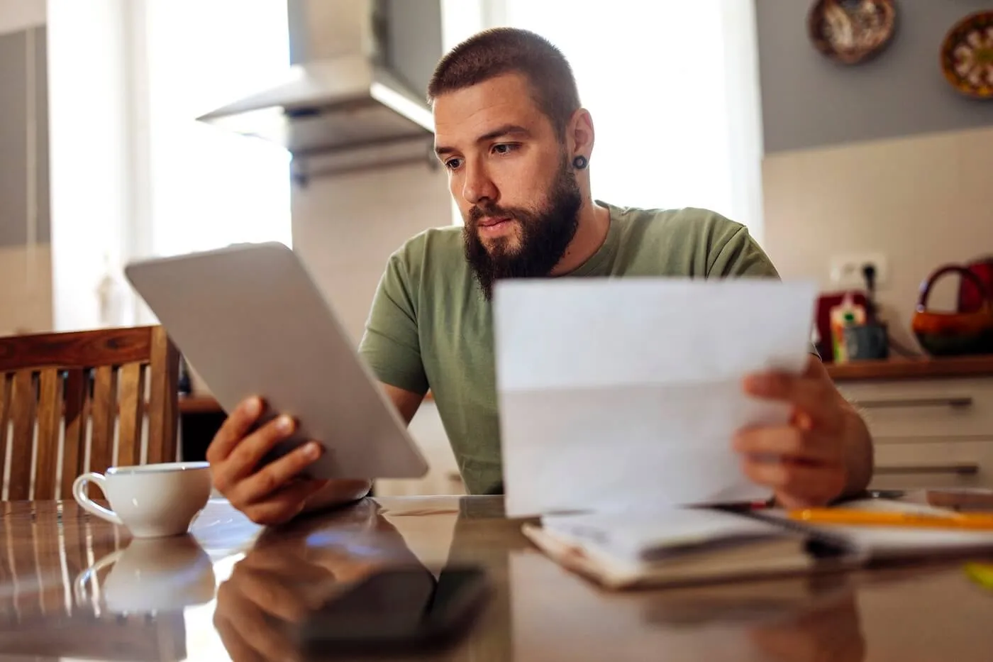 Focused man seated at a kitchen table reviewing a printed document while holding a tablet, with paperwork, a notebook, and a coffee cup arranged on the table in a home setting.
