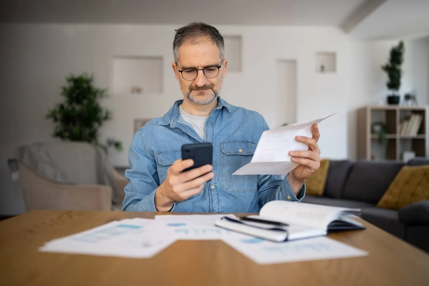 Mature man sitting at a table in a home setting, reviewing paperwork while checking a smartphone, with financial documents, a notebook, and reports spread across the table.