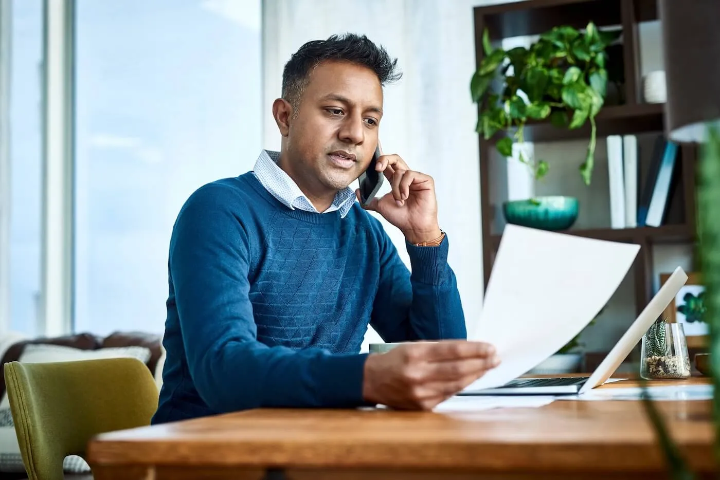 Focused man seated at a wooden table in a home office, holding a printed document while talking on a smartphone. An open laptop, paperwork, and potted plants are visible on and around the workspace, with shelving and large windows in the background.