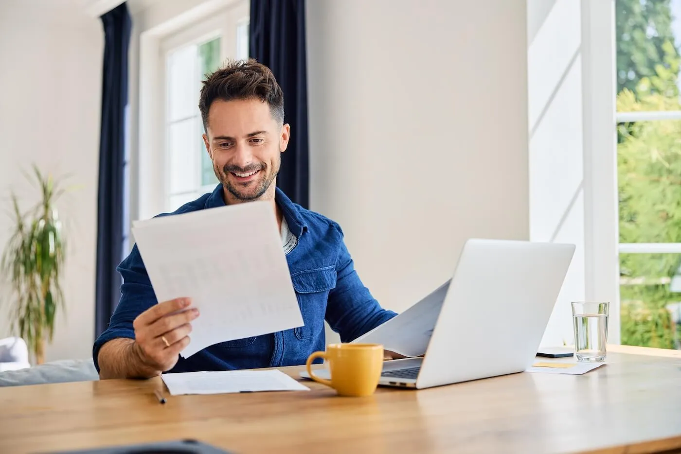 Smiling man seated at a table in a home office reviewing printed documents while using a laptop, with a coffee mug, glass of water, and paperwork arranged on the desk near large windows.