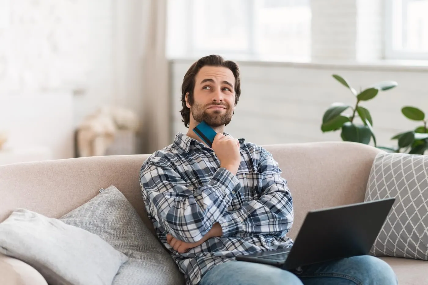 A man sitting on a sofa with a laptop, holding a credit card and looking upward thoughtfully.
