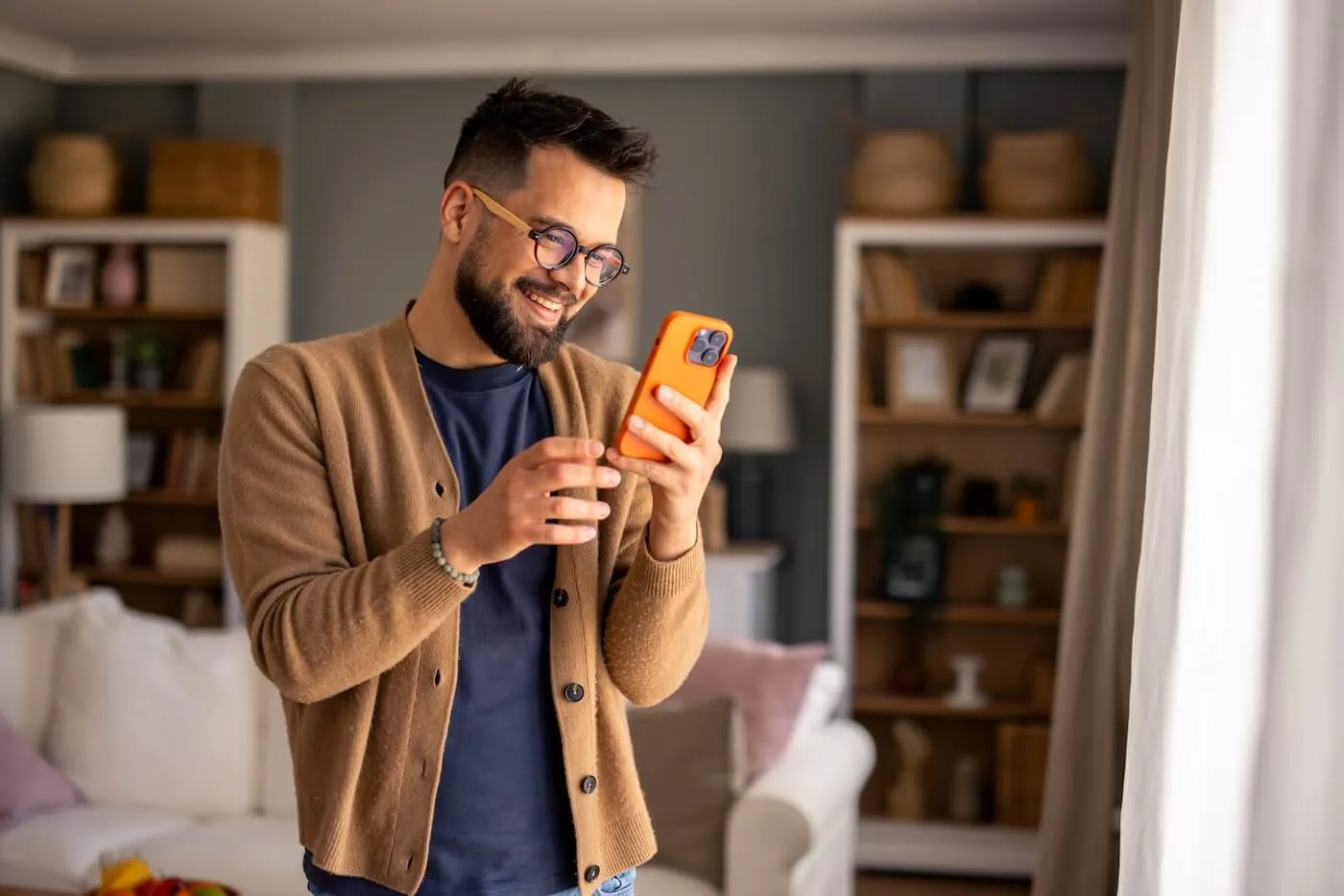 Smiling young man standing in a living room and using a smartphone, with bookshelves, a sofa, and large windows in the background. He is wearing glasses and a cardigan while interacting with a mobile device in a home setting.