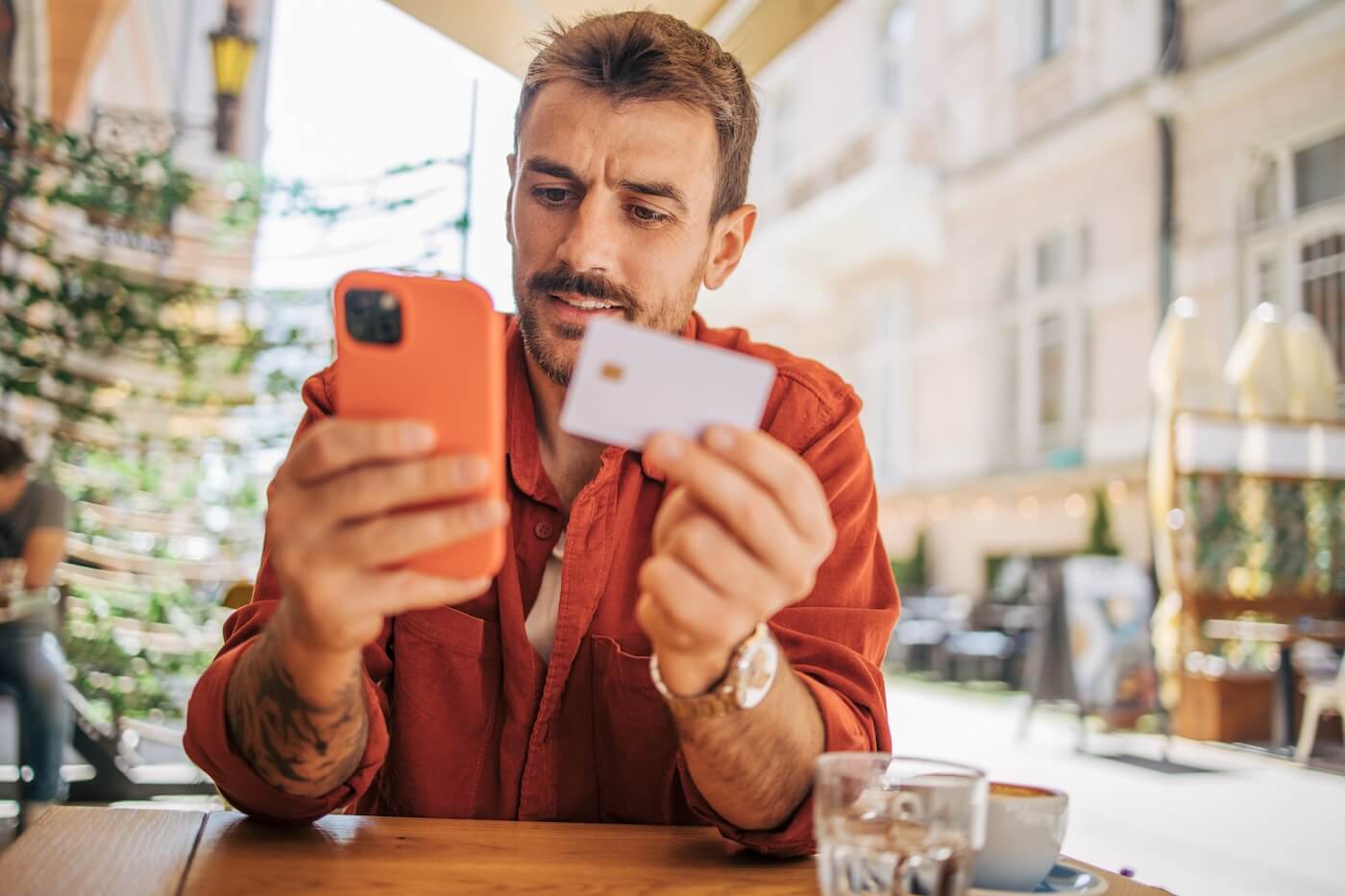 Focused man seated at an outdoor café using a smartphone while holding a credit card at a wooden table with a drink nearby
