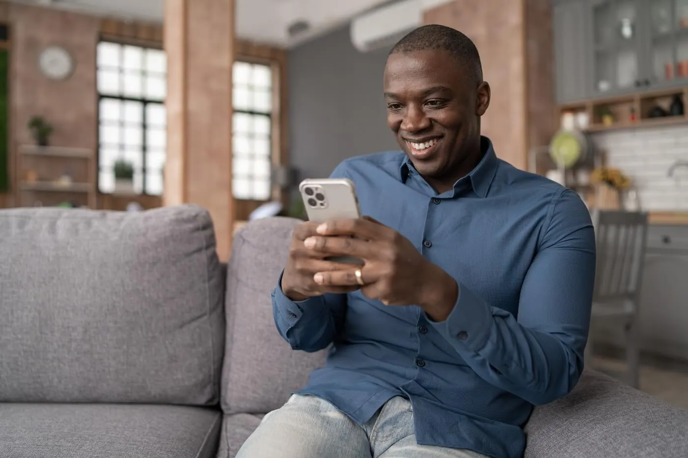 Smiling man seated on a gray sofa in a modern living room, using a smartphone with both hands, with a kitchen area, large windows, and home furnishings visible in the background.