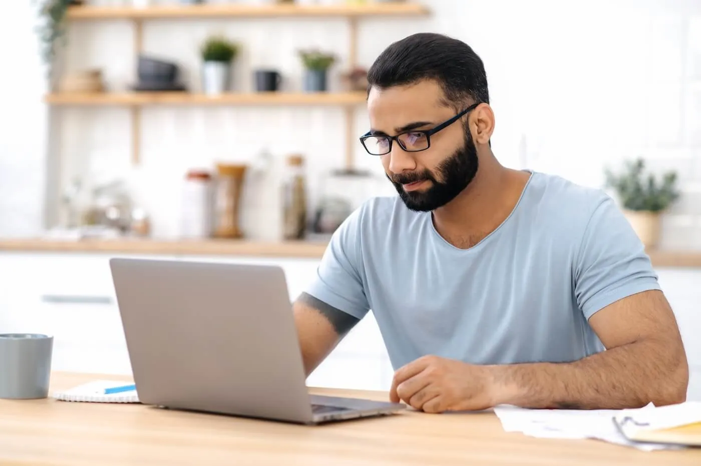 Focused man wearing glasses seated at a kitchen table using a laptop in a bright home setting. A coffee mug, notebook, pen, and papers are on the table, with open shelving, kitchen items, and potted plants visible in the background.