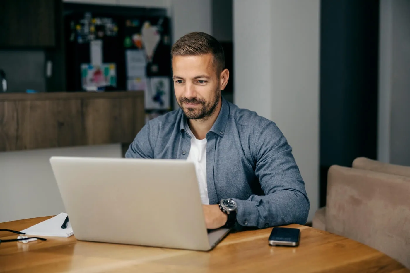 Focused man seated at a wooden table using a laptop in a home setting, with a smartphone, notebook, and pen nearby, and a kitchen area visible in the background.