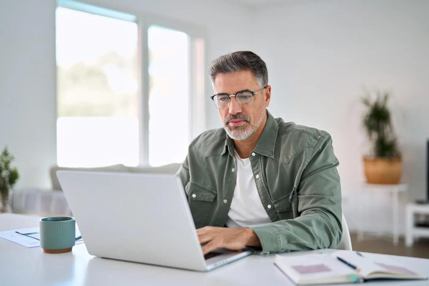 Mature man wearing glasses seated at a desk using a laptop in a bright home office. A coffee mug, notebook, pen, and printed documents are arranged on the desk, with large windows and a potted plant visible in the background.