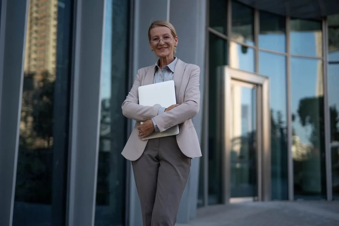 Smiling mature woman standing outside a modern glass office building holding a closed laptop, wearing business attire and positioned near the building entrance with reflective windows in the background.