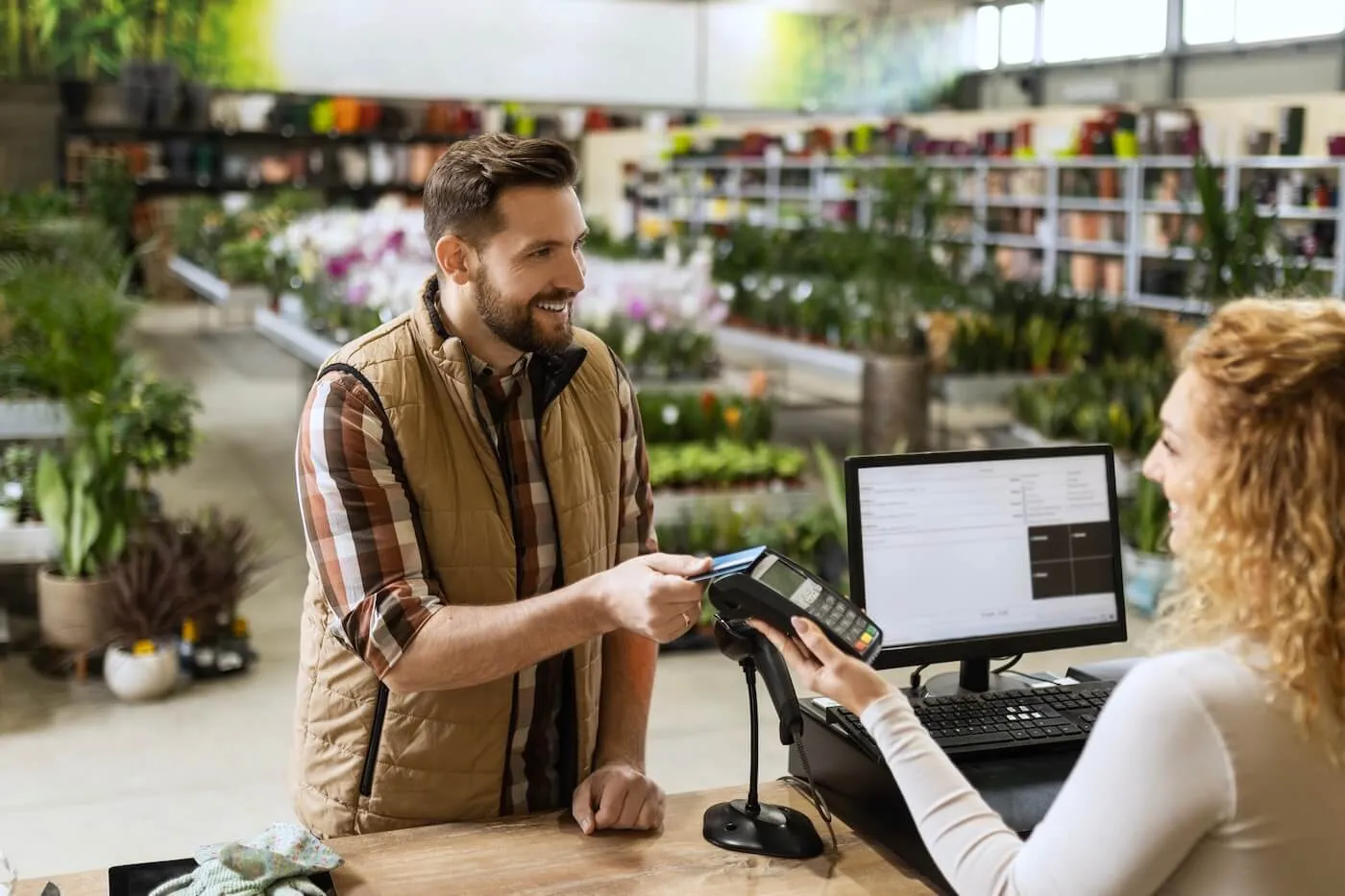 A customer holds a payment card toward a card reader at a counter, while a cashier stands nearby with a computer screen and barcode scanner, inside a store filled with plants and merchandise