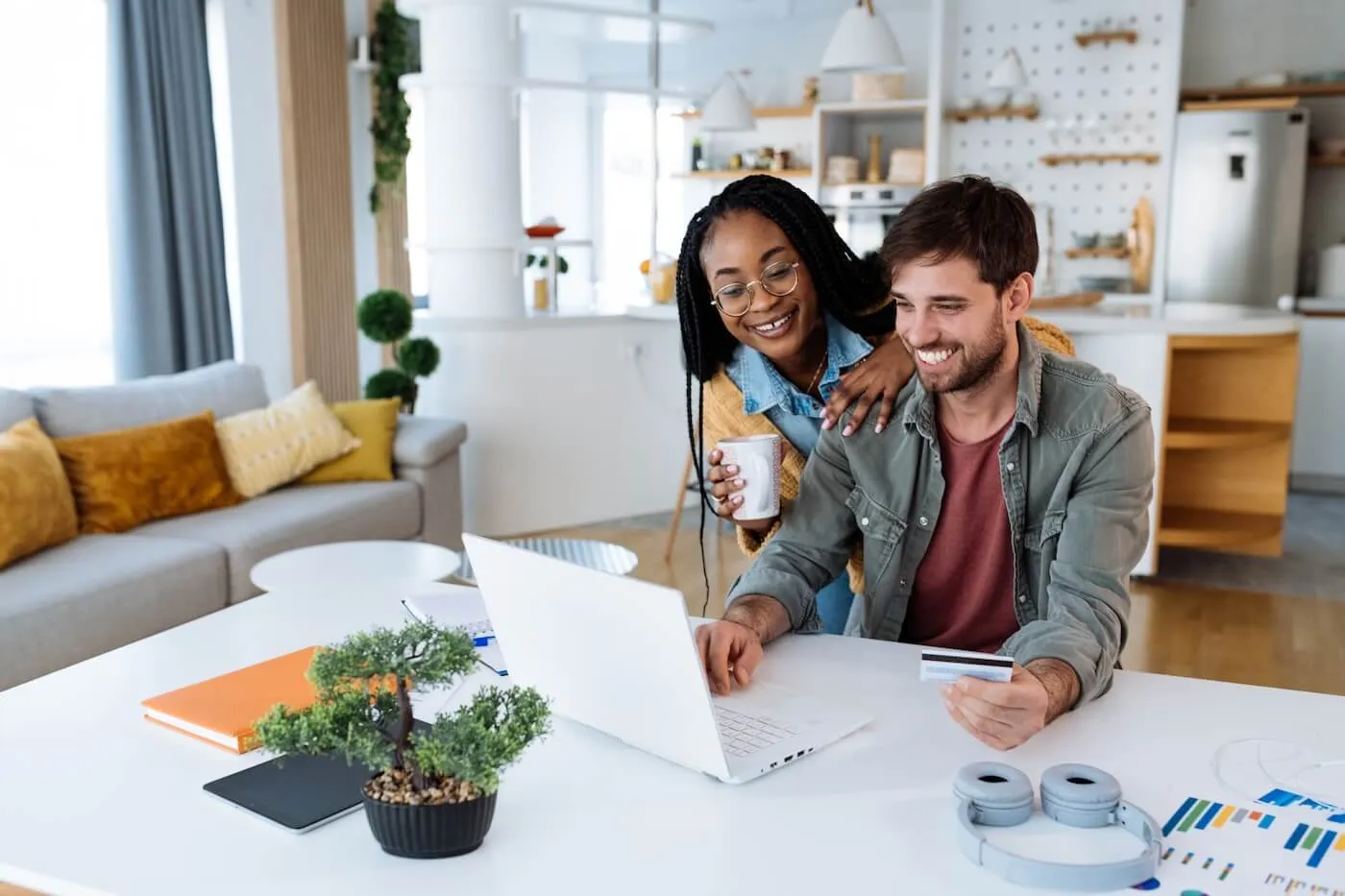 Happy young couple sitting at a home desk with a laptop, using a credit card for an online purchase while enjoying coffee together