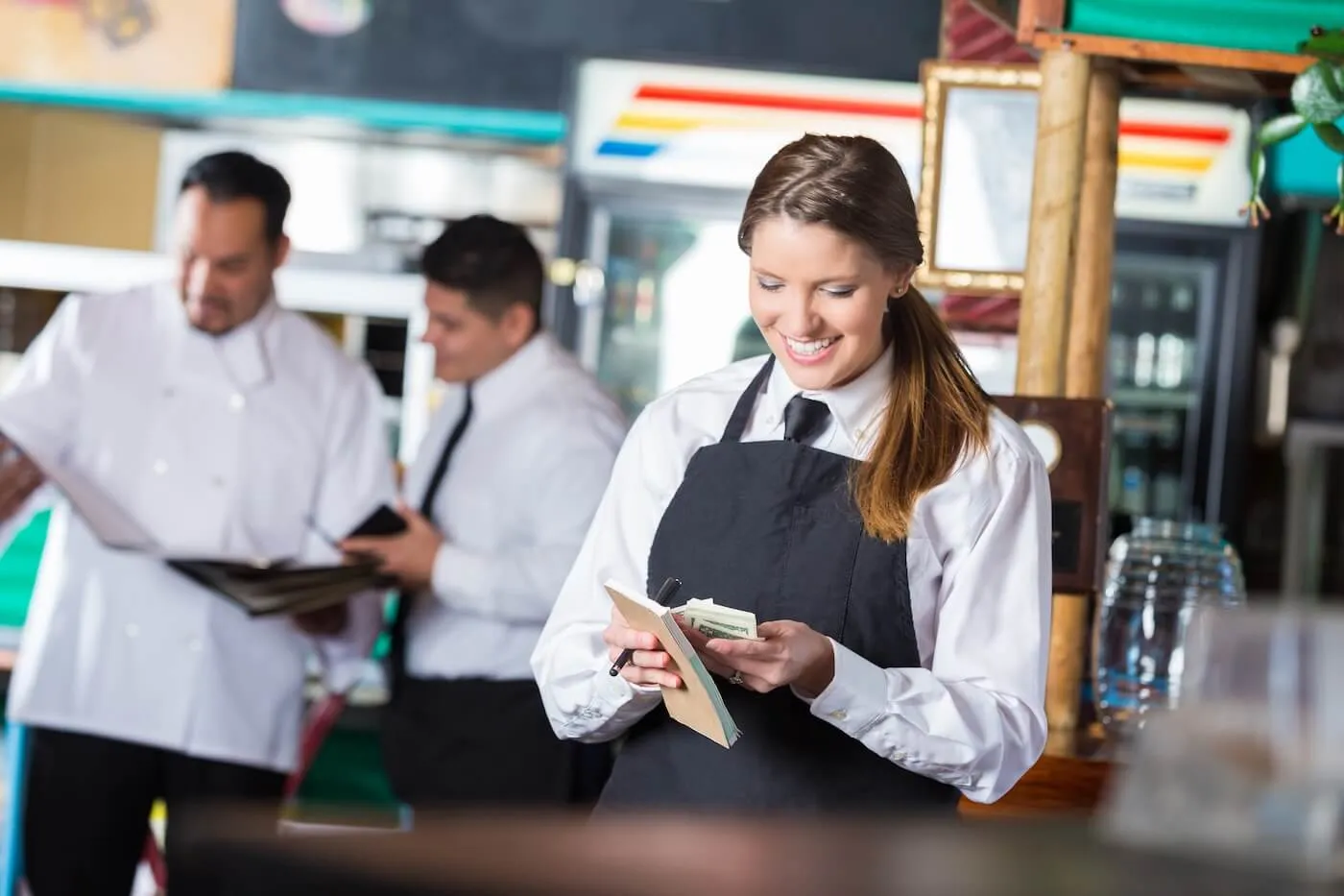 A female restaurant server wearing an apron counts cash with a smile while other staff members work behind her