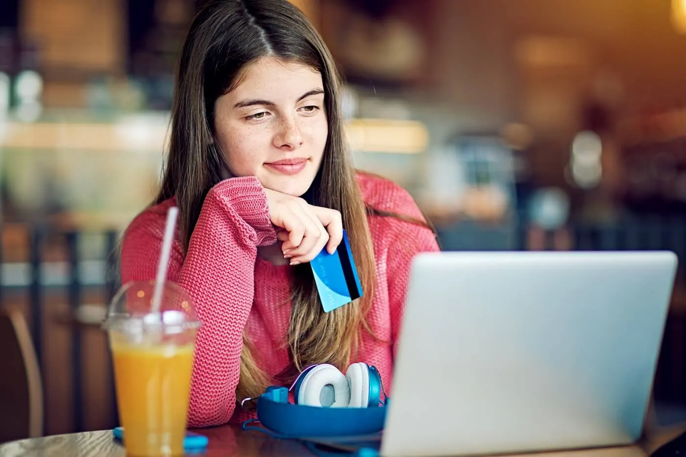 Young woman sitting at a café table with a laptop, holding a credit card, with headphones and a drink beside her