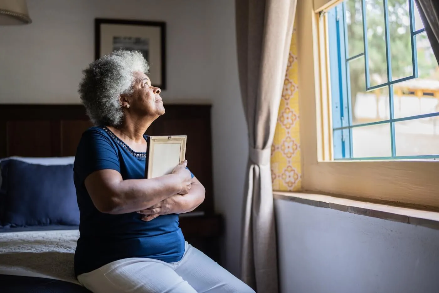Older woman seated beside a bedroom window holding a framed photograph, with natural light streaming through the open window and a bed, curtains, and framed wall art visible in the background.