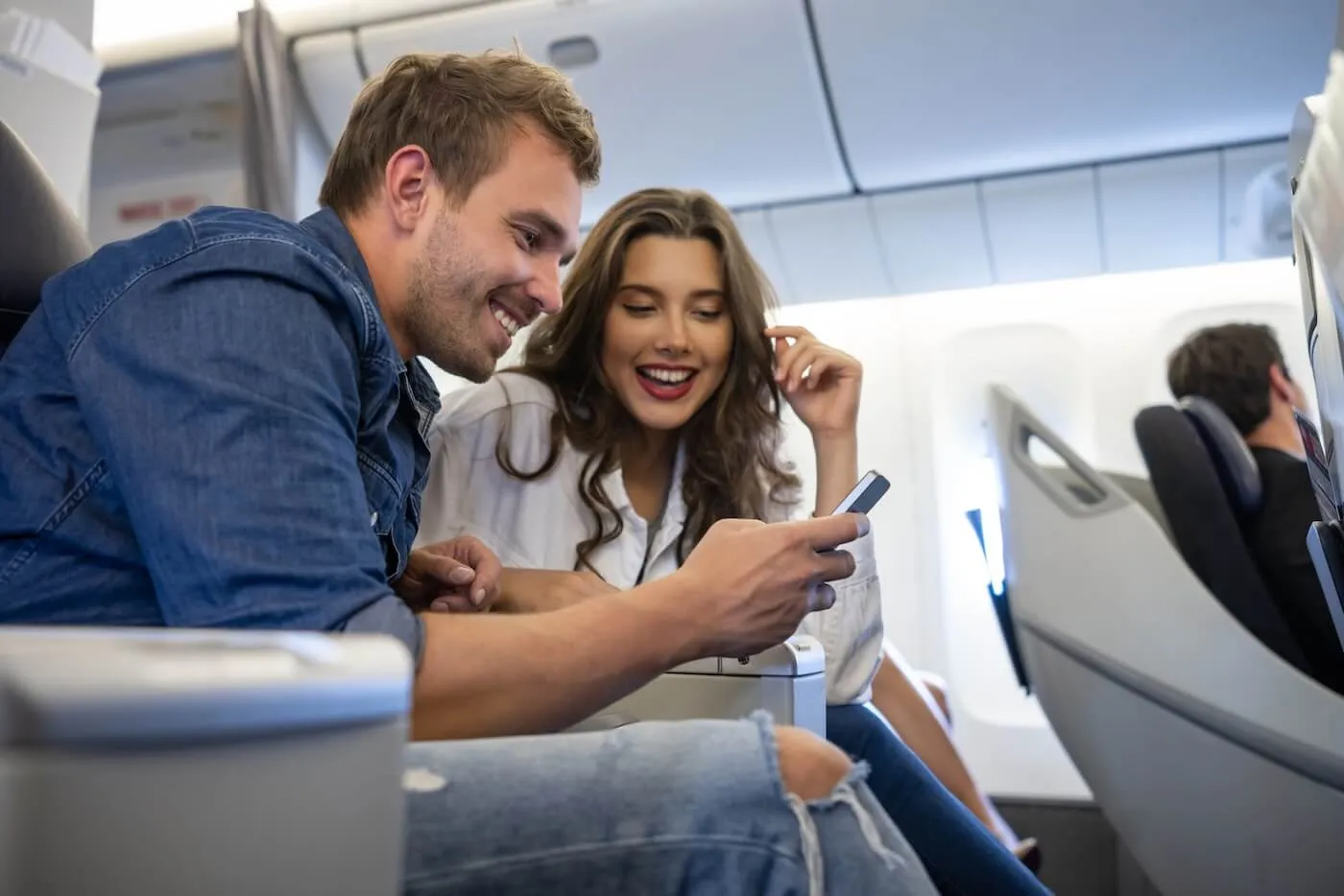 Happy couple seated side by side in a commercial airplane cabin looking at a smartphone together, with aircraft seats, windows, and other travelers visible in the background.