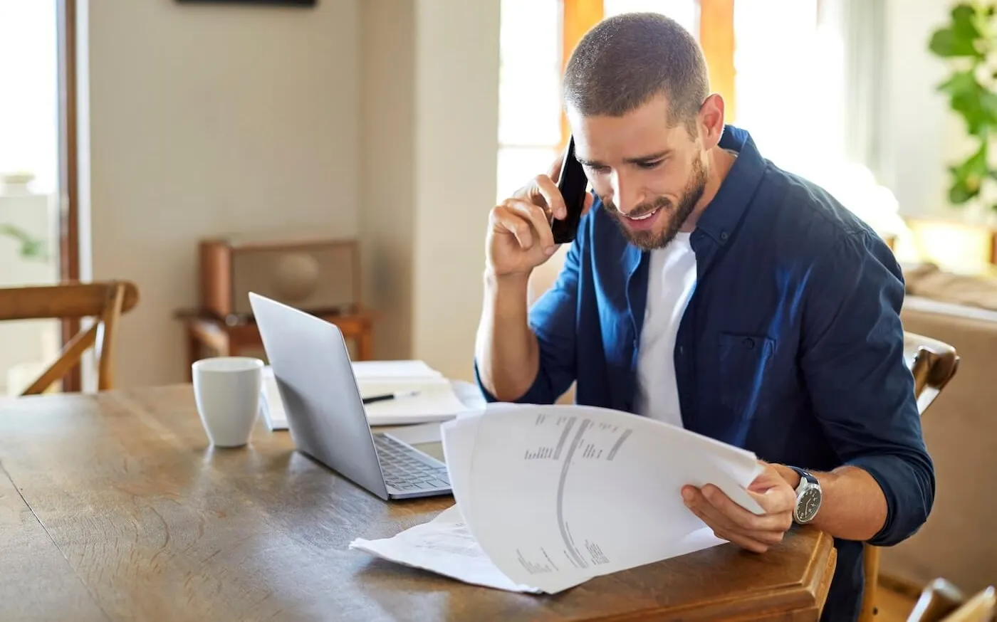 Young man reviews financial documents while speaking on a phone at a table with a laptop and coffee cup