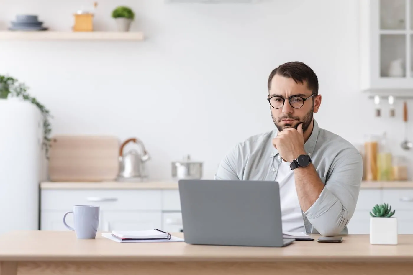 Focused man seated at a desk in a home office or kitchen workspace, looking at a laptop computer, with a notebook, smartphone, coffee mug, and small potted plant on the table.