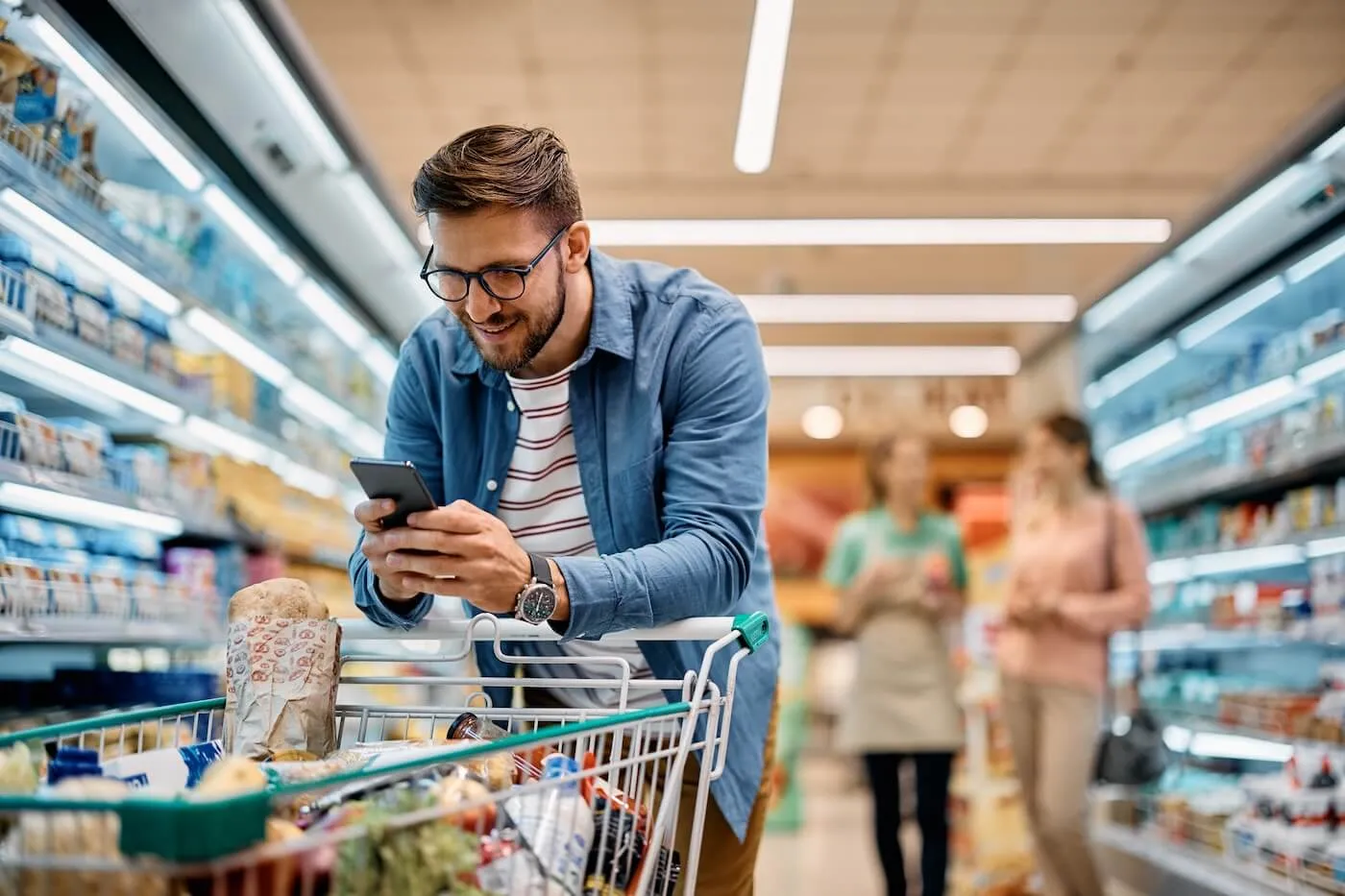 Smiling man wearing glasses pushing a grocery trolley while using his phone in a supermarket, with two women chatting in the background