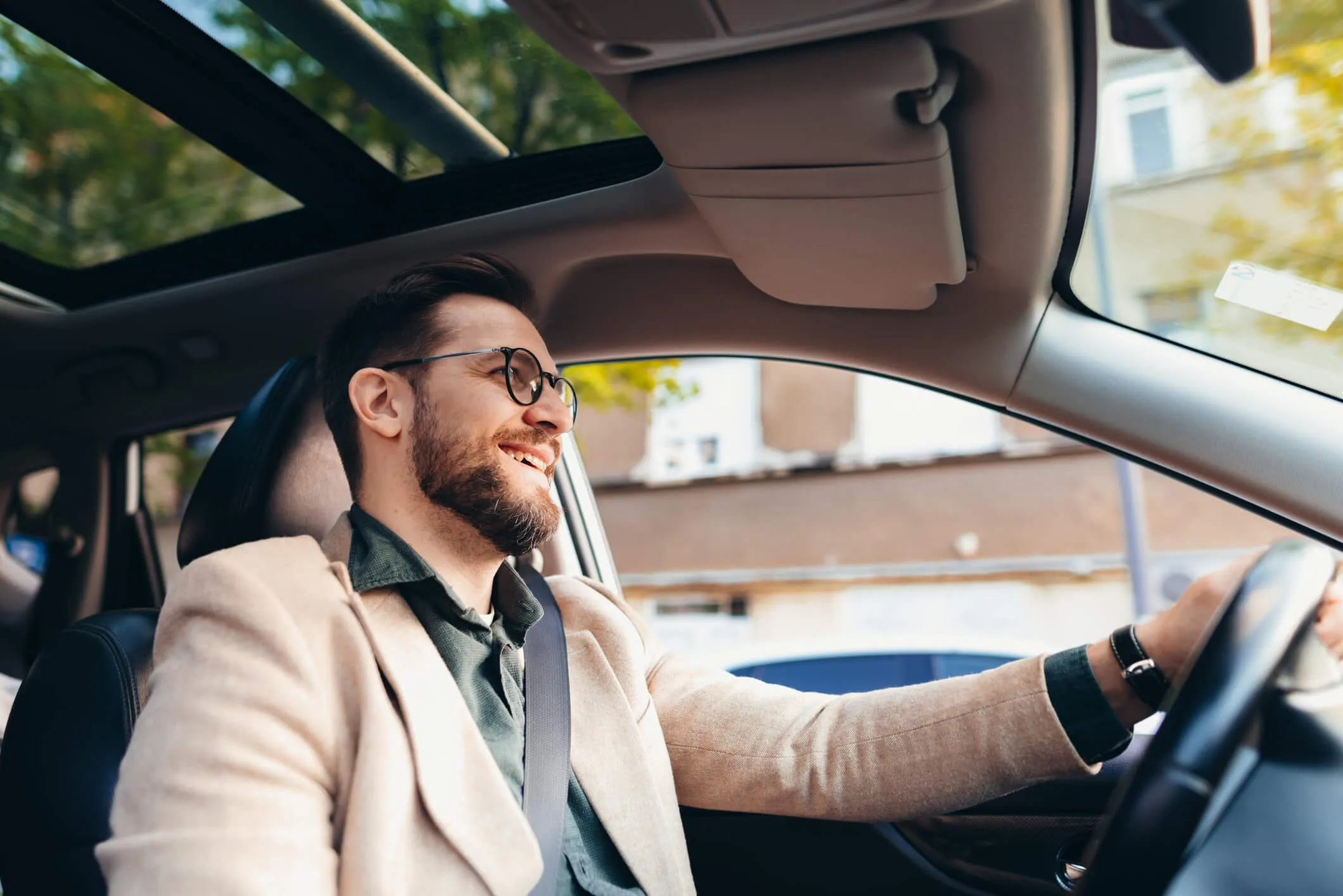 Smiling man sits behind the wheel of a car with sunlight coming through the sunroof while driving through a city street.